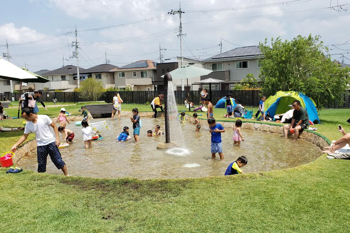 噴水のある浅い水遊び場と、周囲の芝生・日よけのようす。人物の顔はプライバシー保護のためぼかしています